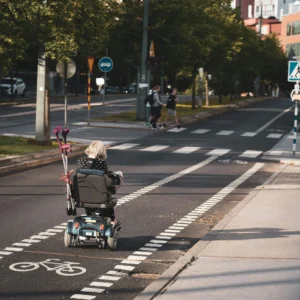 An elderly person riding a mobility scooter on a city street bike lane with crutches attached to the back.