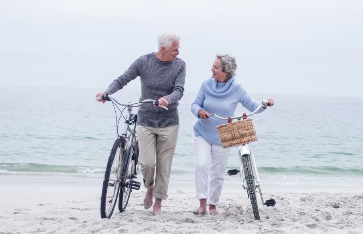 Two elderly people walking on a beach with bicycles