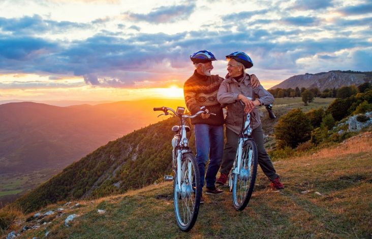 Two elderly people stood together with their bicycles on a hilltop with the sunrise in the background.